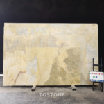 Golden Blue Jade Marble panel in front of stacked slabs, showing cream, taupe and pale blue-gray layered patterns.