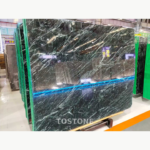 Stacked Greek Verde Alpi marble slabs with fine diagonal white veining across dark green surfaces.