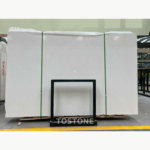 White Sand Beige Marble slab bundle with a pale cream surface, green straps and a black support frame.